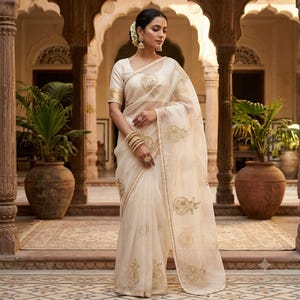 May include: A cream-colored saree featuring intricate gold embroidery in circular patterns throughout the fabric. The garment includes a matching blouse with short sleeves, a sheer pallu draped over the shoulder, and a decorative gold border trim.