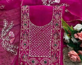 May include: A magenta-colored embroidered salwar kameez set. The top features intricate embroidery with white, green, and gold accents around the neckline and borders. The dupatta has matching floral embroidery. The outfit is displayed on a textured surface.