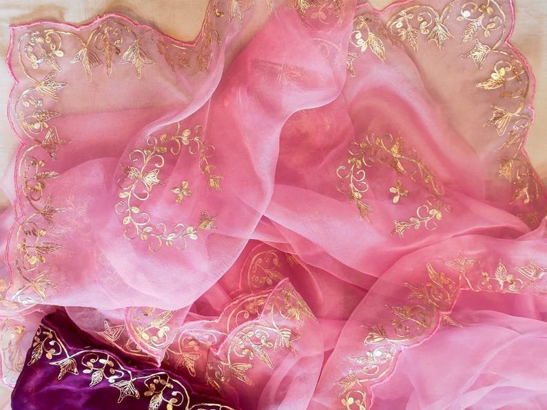 K&ouml;nnte beinhalten: Ein flie&szlig;ender, durchsichtiger rosa Stoff mit aufwendiger goldener Stickerei. Der Stoff ist drapiert und gefaltet und zeigt die zarten Blumen- und Blattmuster. Ein tiefvioletter Stoff mit goldener Stickerei ist unten sichtbar.