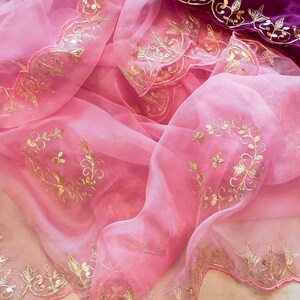 K&ouml;nnte beinhalten: Ein flie&szlig;ender, rosa Stoff mit goldenen Blumenstickereien. Das zarte Material ist drapiert und zeigt die komplizierten goldenen Details an den R&auml;ndern und &uuml;ber die Oberfl&auml;che verteilt. Ein Abschnitt aus tiefviolettem Stoff mit Goldstickerei ist sichtbar.