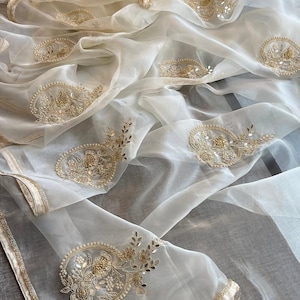 May include: Cream-coloured, sheer fabric with detailed gold embroidery. The fabric is draped, displaying floral and paisley-style patterns. The edges are trimmed with a gold border, adding a touch of elegance. The overall aesthetic is delicate and luxurious.