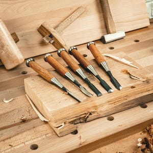Op de afbeelding: Een verzameling houtbewerkingsgereedschap, waaronder vijf beitels met houten handvatten en metalen bladen, gerangschikt op een houten oppervlak. Een houten hamer en ander gereedschap zijn ook zichtbaar, wat wijst op een timmer- of houtbewerkingsthema.