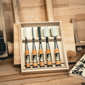 Peut inclure: Un ensemble de cinq ciseaux à bois avec des manches en bois et des lames en métal noir, présentés dans une boîte en bois. Les ciseaux ont un design traditionnel avec une bande noire et du texte japonais sur les manches. L'arrière-plan comprend des planches de bois et une surface de travail en bois.