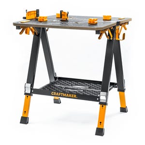 Op de afbeelding: Een zwart-gele draagbare werkbank met een gedeeld tafelblad, verstelbare oranje klemblokken en een onderste opbergplank. Het frame heeft metalen poten met hoogteverstellingsmarkeringen en de tekst CRAFTMAKER op de voorzijde.