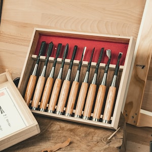Może przedstawiać: Zestaw dłut do rzeźbienia w drewnie w drewnianym pudełku. Dłuta mają drewniane uchwyty, srebrne metalowe tuleje i czarne ostrza. Pudełko jest wyłożone czerwoną tkaniną i stoi na drewnianej powierzchni.