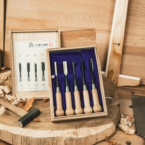 Puede incluir: Un juego de cinco cinceles para tallar madera con mangos de madera y hojas de metal, presentados en una caja de madera con interior azul. Una segunda caja de madera contiene un diagrama de los cinceles. Los cinceles están dispuestos sobre una superficie de madera.