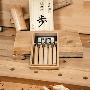 Könnte beinhalten: Ein Satz von fünf Holzbearbeitungswerkzeugen mit Holzgriffen, präsentiert in einer Holzkiste. Die Kiste befindet sich in einer größeren Holzkiste mit japanischem Text. Ein Holzhammer und andere Holzbearbeitungswerkzeuge sind ebenfalls sichtbar.