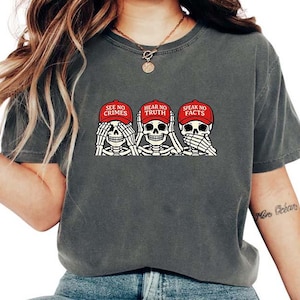 May include: Dark gray t-shirt featuring a graphic of three skeleton heads, each with hands covering their eyes, ears, and mouth. The skeletons wear red caps with the phrases "SEE NO CRIMES", "HEAR NO TRUTH", and "SPEAK NO FACTS".