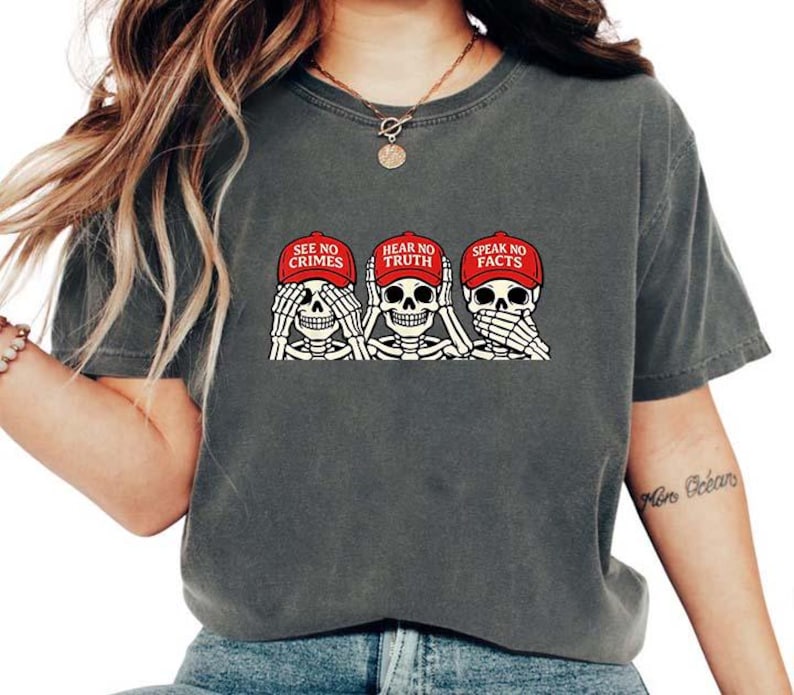 May include: Dark gray t-shirt featuring a graphic of three skeleton heads, each with hands covering their eyes, ears, and mouth. The skeletons wear red caps with the phrases "SEE NO CRIMES", "HEAR NO TRUTH", and "SPEAK NO FACTS".