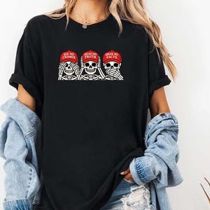 May include: Black t-shirt featuring a graphic of three skeleton figures, each with hands covering their eyes, ears, and mouth. The skeletons wear red caps with the phrases "SEE NO CRIMES", "HEAR NO TRUTH", and "SPEAK NO FACTS".