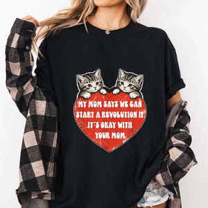 May include: Black t-shirt featuring two gray tabby kittens peeking over a red heart. The heart contains the text "MY MOM SAYS WE CAN START A REVOLUTION IF IT'S OKAY WITH YOUR MOM" in white letters.
