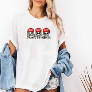 May include: White t-shirt featuring a graphic of three skeleton heads wearing red caps. The caps have the phrases "SEE NO CRIMES", "HEAR NO TRUTH", and "SPEAK NO FACTS". The design is in black and red.