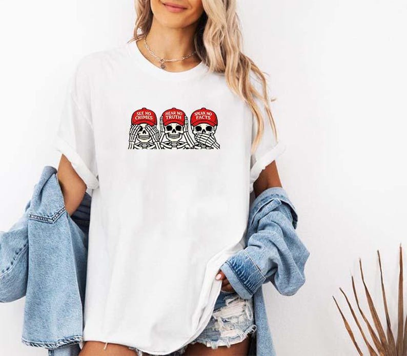 May include: White t-shirt featuring a graphic of three skeleton heads wearing red caps. The caps have the phrases "SEE NO CRIMES", "HEAR NO TRUTH", and "SPEAK NO FACTS". The design is in black and red.