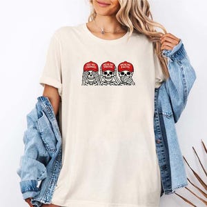 May include: Cream-colored t-shirt featuring a graphic of three skeleton heads, each wearing a red cap. The caps read "SEE NO CRIMES", "HEAR NO TRUTH", and "SPEAK NO FACTS". The design is in black and red.
