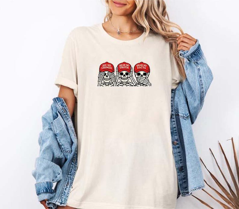 May include: Cream-colored t-shirt featuring a graphic of three skeleton heads, each wearing a red cap. The caps read "SEE NO CRIMES", "HEAR NO TRUTH", and "SPEAK NO FACTS". The design is in black and red.