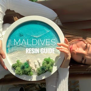 Könnte beinhalten: Rundes, weiß gerahmtes Kunstwerk aus Kunstharz mit dem Thema Malediven. Das Kunstwerk zeigt einen türkisfarbenen Ozean, weißen Sand und grünes Moos. Der Text "MALDIVES RESIN GUIDE" ist in Weiß dargestellt.