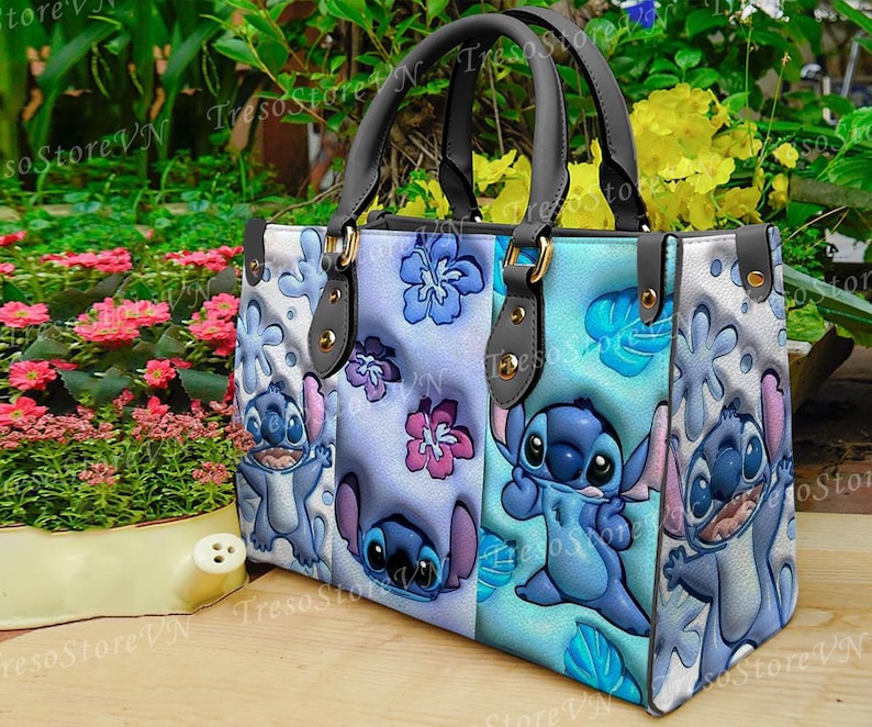 K&ouml;nnte beinhalten: Eine Tragetasche mit Stitch-Design aus Lilo & Stitch. Die Tasche hat einen hellblauen Hintergrund mit Stitch, Blumen und Bl&auml;ttermustern. Sie hat schwarze Griffe und goldfarbene Beschl&auml;ge. Ein farbenfrohes Accessoire.