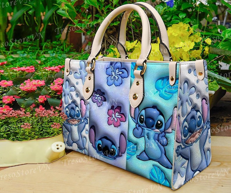 K&ouml;nnte beinhalten: Eine Tragetasche mit einem farbenfrohen Design mit der Figur Stitch aus Lilo & Stitch. Die Tasche hat ein hellblaues, lila und wei&szlig;es Farbschema mit Stitch-Illustrationen und floralen Akzenten. Sie hat cremefarbene Griffe und goldfarbene Hardware.