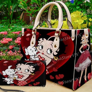 Pode incluir: Uma bolsa preta e uma carteira a condizer com uma imagem de desenho animado de Betty Boop com um cão branco e corações vermelhos. A mala tem alças e acabamentos creme. A carteira tem uma alça de pulso.