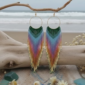 May include: A pair of long, beaded earrings with a gradient of colors, including green, blue, purple, orange, and yellow. The earrings are suspended from gold-colored hoops and are displayed against a beach backdrop with driftwood and ocean waves.