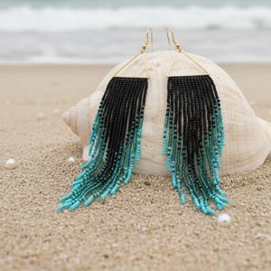 May include: A pair of beaded earrings with a black and turquoise ombre design. The earrings have a gold-colored triangular top and long fringe. The earrings are displayed on a seashell on a sandy beach.