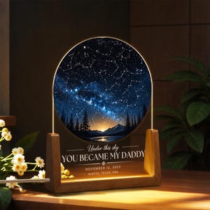 Könnte beinhalten: Ein Nachtlicht mit Holzsockel und einem kreisförmigen, beleuchteten Panel, das einen Sternenhimmel zeigt. Das Panel zeigt eine Sternenkarte, eine Bergkette und den Text "Under this sky YOU BECAME MY DADDY" mit Datum und Ort.