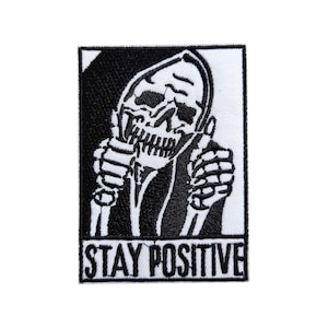 May include: Black and white embroidered patch featuring a skeleton giving a thumbs up. The skeleton is wearing a hooded robe. The text "STAY POSITIVE" is at the bottom of the patch. The patch is rectangular.