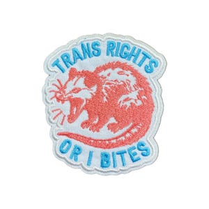 May include: Embroidered patch with the text "TRANS RIGHTS OR I BITES" in light blue, surrounding a coral-colored opossum illustration. The patch has a white border and is designed for application on clothing or accessories.