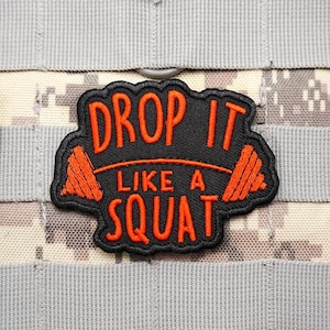 May include: A black patch with orange text that reads "DROP IT LIKE A SQUAT". The patch has an orange barbell design on each side of the text. The patch is on a camouflage background.