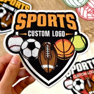 May include: Embroidered patch with the word "SPORTS" in orange, and "CUSTOM LOGO" in white. The patch features a baseball, soccer ball, football, basketball, and tennis ball. The patch is black with a white border.