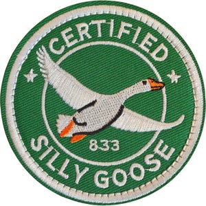 Puede incluir: Parche bordado circular con fondo verde y borde blanco. Presenta un ganso volando con pico y patas naranjas. El texto "CERTIFIED SILLY GOOSE" está en círculo, con el número "833" también incluido.