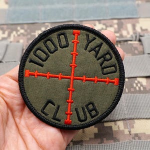 May include: A round, olive green patch with the text "1000 YARD CLUB" in black. A red crosshair design is centered on the patch. The patch has a black border and is likely an emblem for a shooting or military organization.