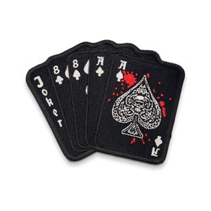 May include: A set of black playing card patches. The cards include a Joker, two 8s of spades, and an Ace of Spades with a skull design and red splatter. The patches are embroidered with white details.
