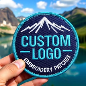May include: A round, navy blue embroidered patch with a mountain design in white thread. The text "CUSTOM LOGO" and "EMBROIDERY PATCHES" are in light blue and white. The patch has a light blue border.
