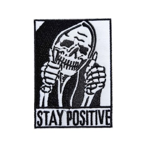 May include: A black and white embroidered patch featuring a skeleton with a thumbs-up gesture. The skeleton is wearing a hooded robe. The text "STAY POSITIVE" is displayed in a bold, sans-serif font at the bottom of the patch.