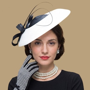 May include: A white hat with a black and white bow and feather accents. The hat is paired with a black and white houndstooth glove and a triple strand pearl necklace. The woman is wearing pearl earrings and a black dress.