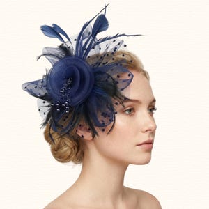 Könnte beinhalten: Ein marineblauer Fascinator mit Blumenmuster, Federn und gepunktetem Netz. Das Kopfstück hat eine zentrale Stoffblume, die mit Federn und einer Perlenverzierung verziert ist. Ein stilvolles Accessoire für besondere Anlässe.
