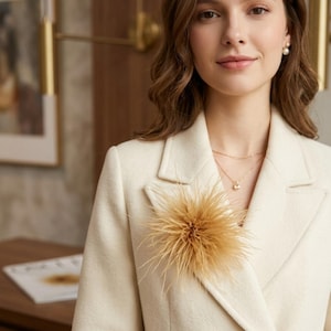 May include: A cream-colored blazer with a double-breasted design. A decorative, fluffy, tan-colored feather brooch is pinned to the lapel. The person is wearing a gold necklace and pearl earrings.