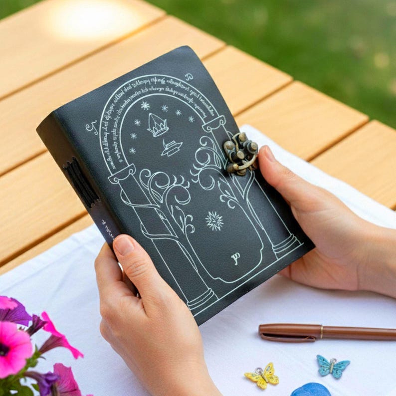 Puede incluir: Un diario de cuero negro con una ilustraci&oacute;n blanca y un cierre de metal. La cubierta presenta un dise&ntilde;o arqueado con una corona y un motivo de &aacute;rbol. El diario est&aacute; abierto, revelando las p&aacute;ginas interiores.