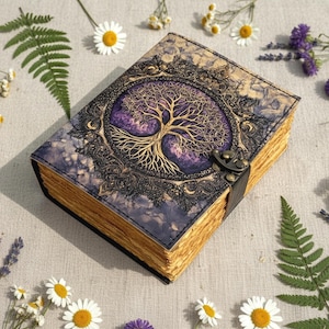 May include: A leather-bound journal with a tree of life design. The cover features a gold-colored tree against a purple background, framed by an ornate border. The journal has a clasp closure and aged-looking pages. Daisies and ferns surround the journal.