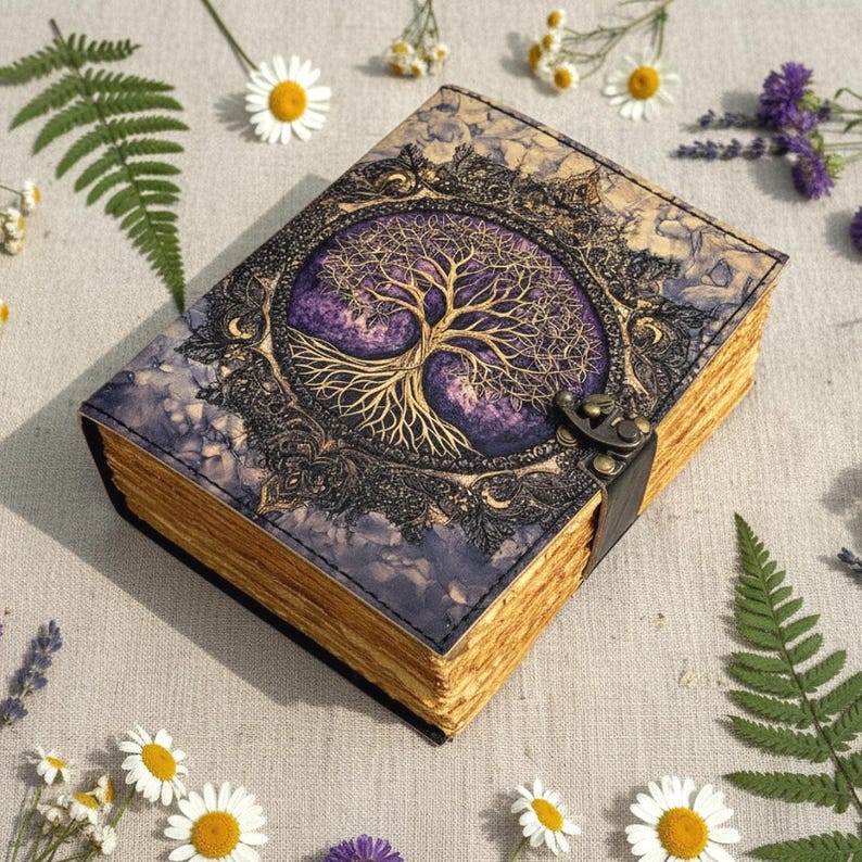 May include: A leather-bound journal with a tree of life design. The cover features a gold-colored tree against a purple background, framed by an ornate border. The journal has a clasp closure and aged-looking pages. Daisies and ferns surround the journal.