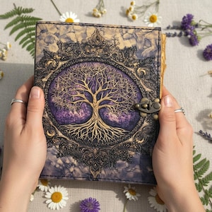May include: A leather-bound journal with a Tree of Life design. The cover features a purple and gold tree illustration within an ornate frame. The journal is held open, showcasing the detailed artwork and clasp closure.