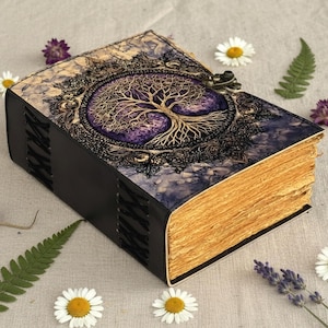 May include: A handcrafted journal with a tree of life design. The cover features a gold-colored tree on a purple background, framed by ornate detailing. The book has a black spine and aged-looking pages, secured with a metal clasp. The journal is surrounded by flowers and greenery.