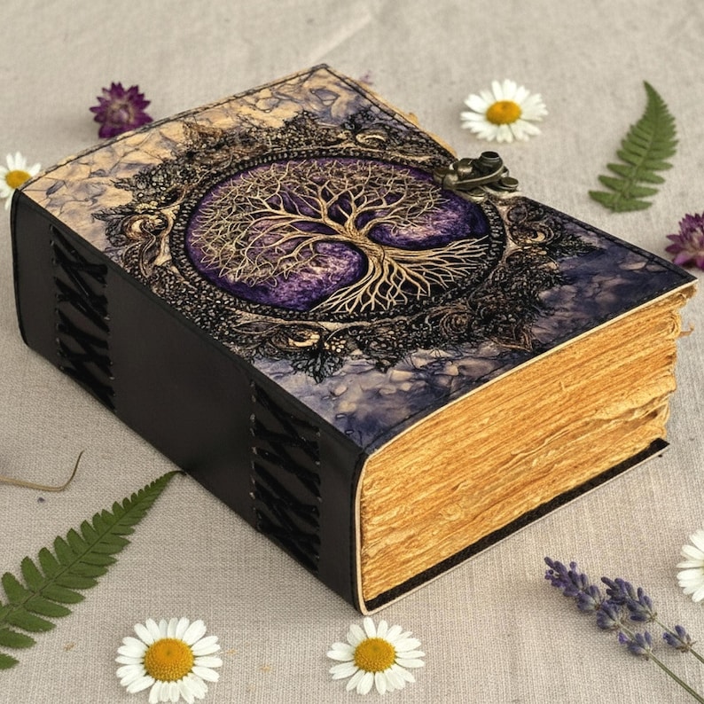 May include: A handcrafted journal with a tree of life design. The cover features a gold-colored tree on a purple background, framed by ornate detailing. The book has a black spine and aged-looking pages, secured with a metal clasp. The journal is surrounded by flowers and greenery.