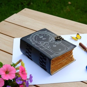 Puede incluir: Diario de cuero negro con un dise&ntilde;o decorativo y un cierre de metal. El diario tiene una encuadernaci&oacute;n cosida y p&aacute;ginas de papel envejecido. Un bol&iacute;grafo y una mariposa amarilla est&aacute;n junto al diario sobre una superficie blanca.