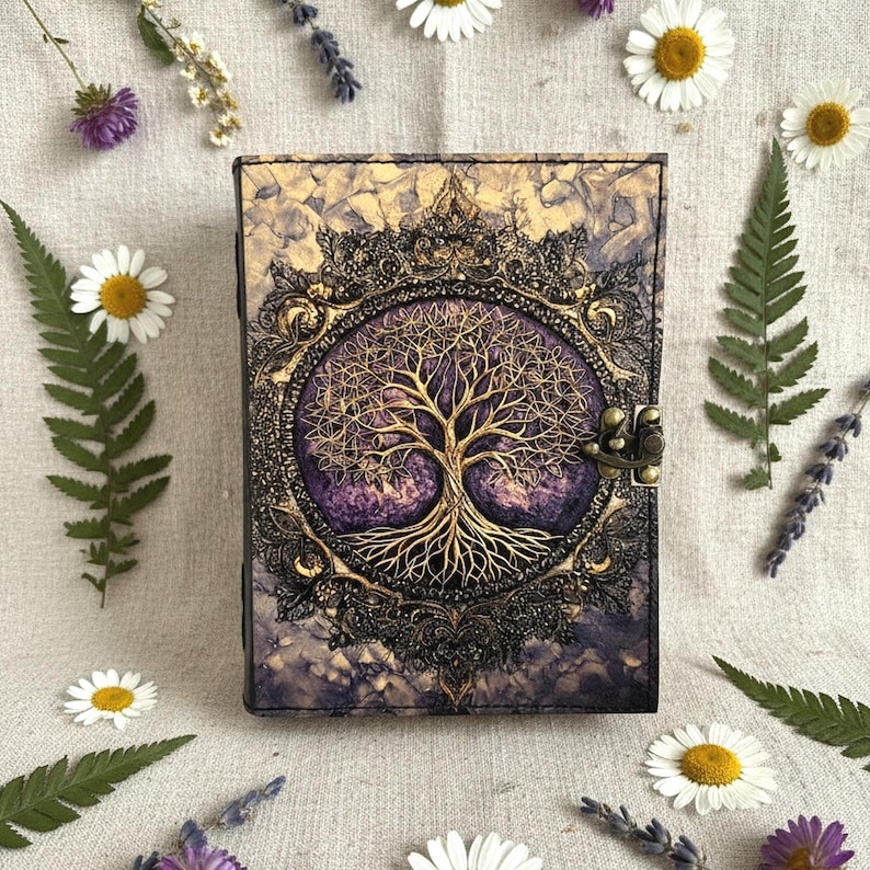 May include: A decorative journal with a gold tree of life design on a purple background. The cover is framed by an ornate, dark bronze border and a bronze clasp. The journal is surrounded by white daisies, ferns, and lavender sprigs.