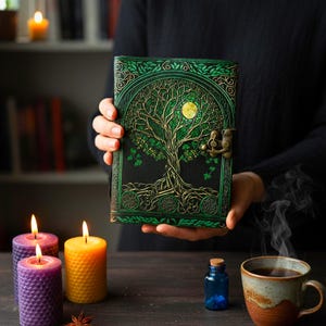 May include: A green and gold embossed journal with a tree design and a full moon. The journal is held in front of a dark background with lit candles, a cup of tea, and a small blue bottle.