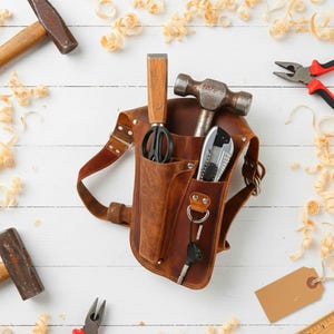 May include: A brown leather tool belt with various tools, including a hammer, chisel, utility knife, and pliers. The belt is surrounded by wood shavings and additional hammers. A key hangs from the belt.