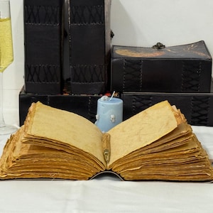 Pu&ograve; includere: Un libro aperto con pagine ingiallite e una penna. Dietro, scatole e libri rilegati in pelle nera e una candela azzurra. A sinistra, un bicchiere di champagne. Suggerisce un tema vintage o antico.