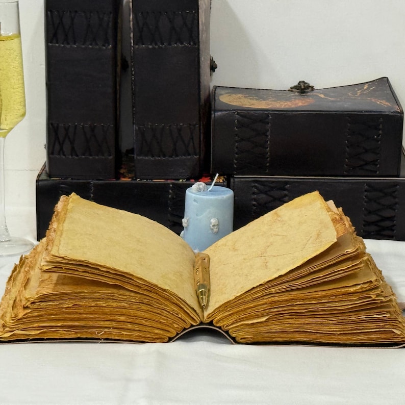 Pu&ograve; includere: Un libro aperto con pagine ingiallite e una penna. Dietro, scatole e libri rilegati in pelle nera e una candela azzurra. A sinistra, un bicchiere di champagne. Suggerisce un tema vintage o antico.